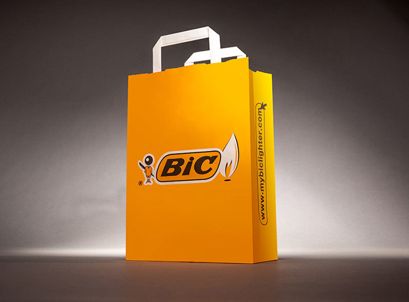 Papiertragetasche mit flachen Henkeln in weiß. BiC als Motiv in Gelb auf der Vorderseite und seitlichem Druck